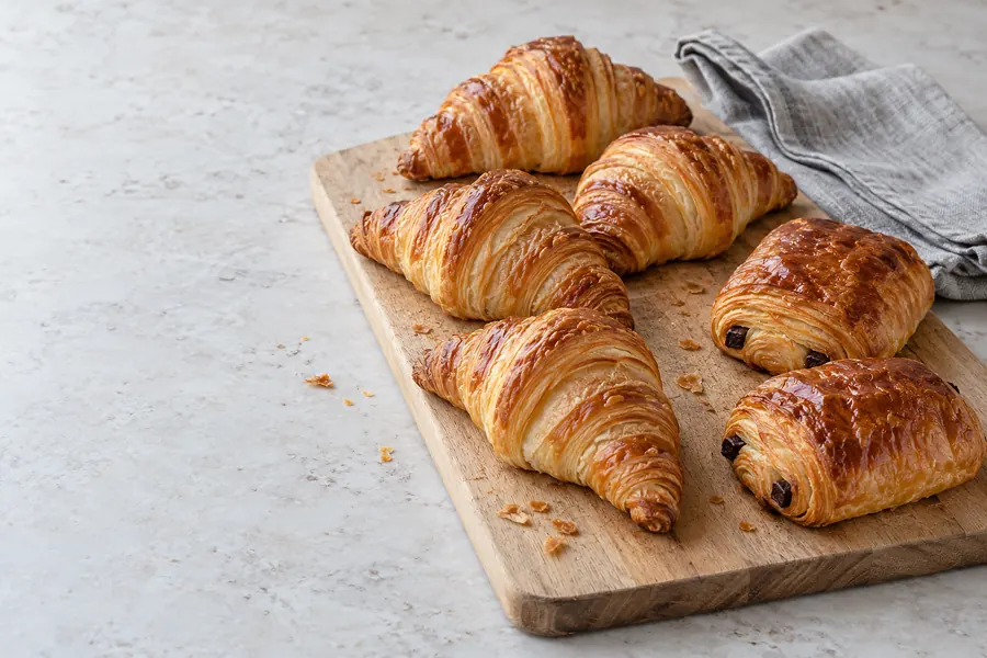 Vier Butter-Croissants und zwei Pain au Chocolat auf einem hellen Holzbrett, daneben eine graue Leinenserviette