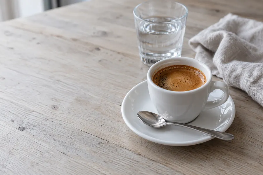Espresso mit dichter Crema in einer kleinen weißen Tasse auf einer Untertasse, dahinter ein Glas Wasser