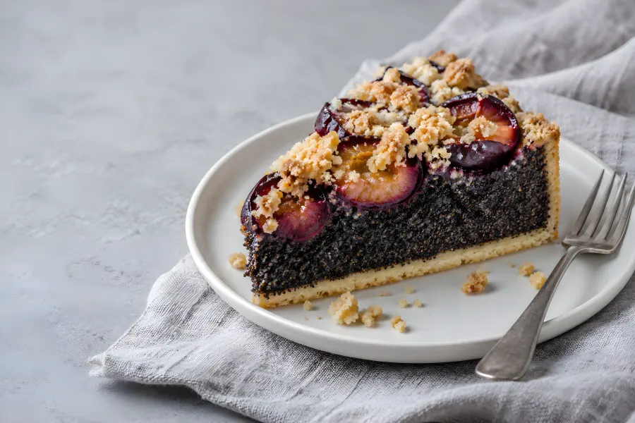 Ein Stück Mohnkuchen mit halbierten Zwetschgen und Streuseln auf einem kleinen weißen Teller mit Gabel