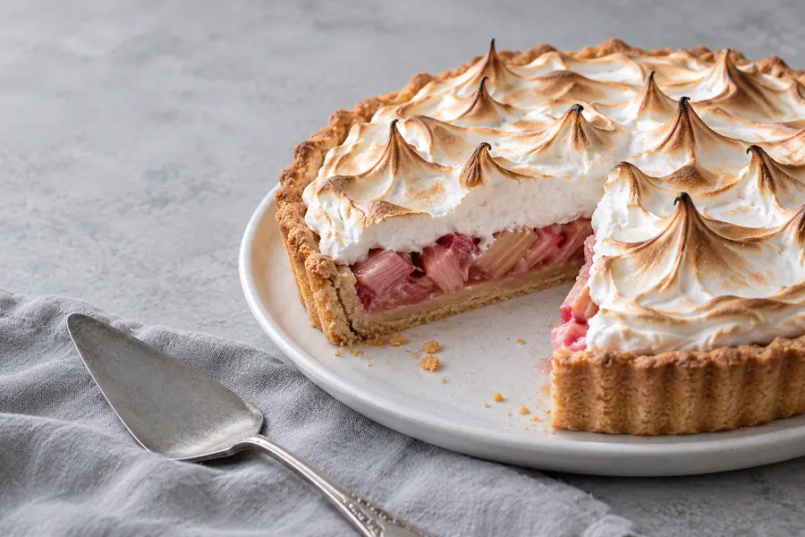 Rhabarber-Baiser-Tarte auf einem weißen Teller, ein Stück herausgeschnitten, daneben ein Tortenheber auf grauem Leinen